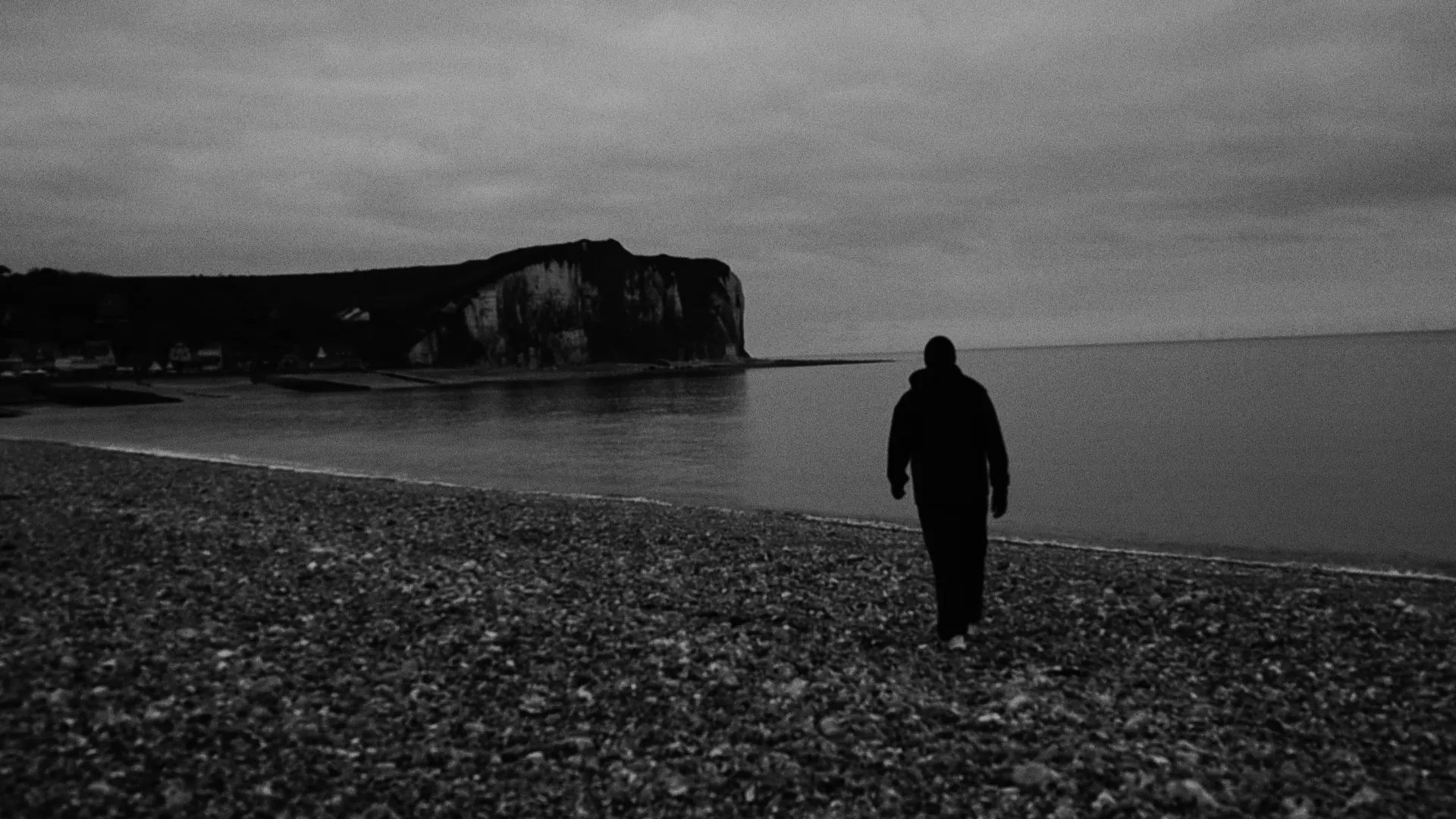 Silhouette solitaire marchant sur une plage de galets face aux falaises, noir et blanc
