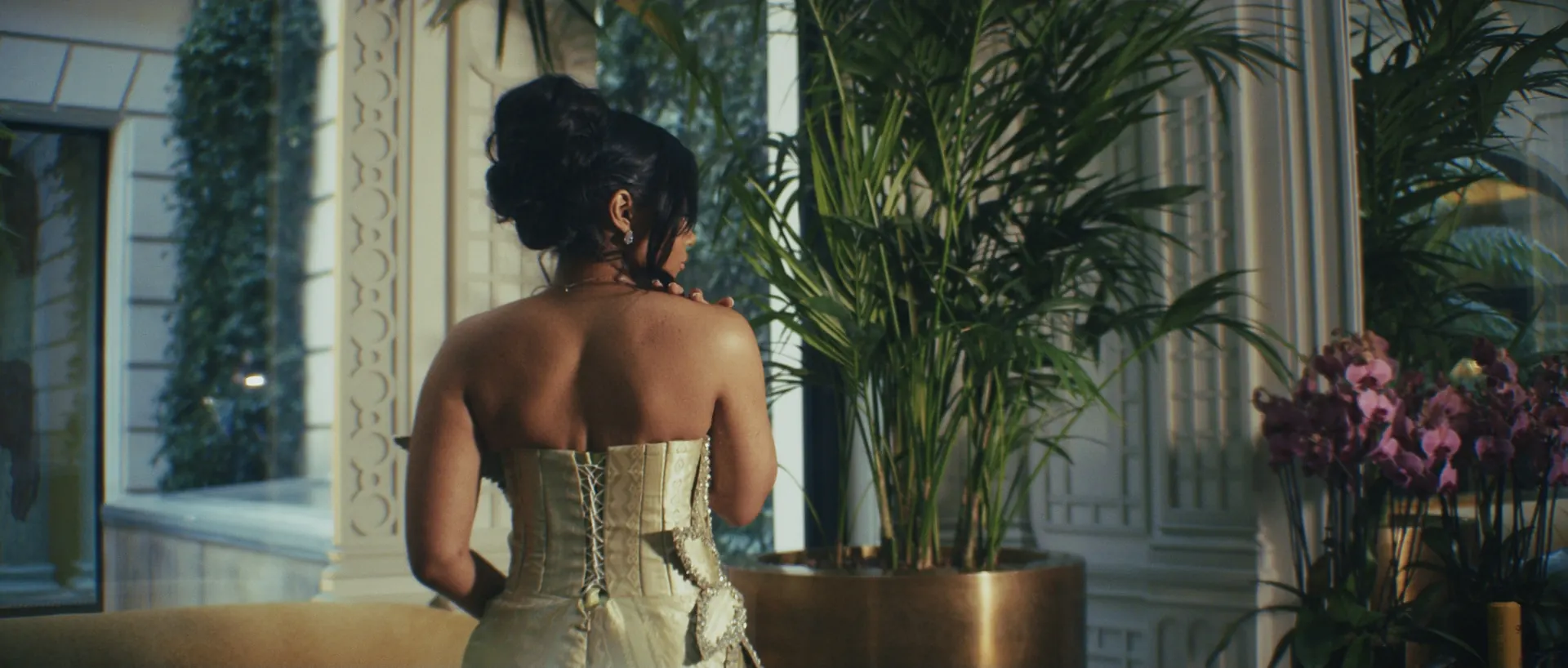 Salon lumineux aux grandes plantes et bouquets, une femme en robe claire de dos